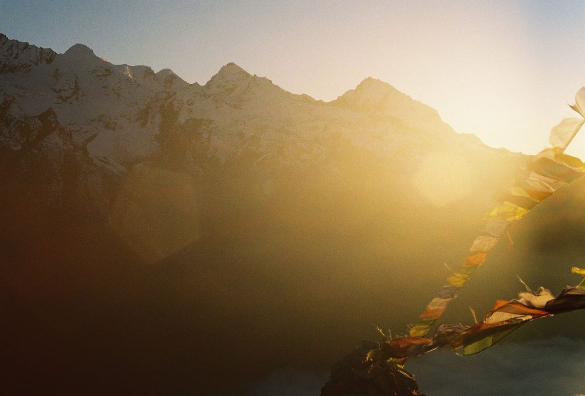 Mountain ridges in near-silhouette under an intense sun flare, prayer flags crossing the lower right.