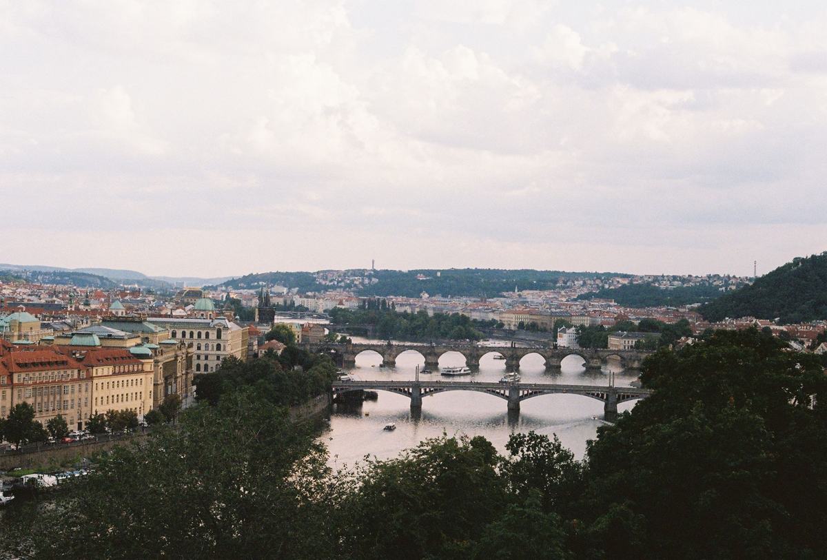 A European city seen from above its river, a stone bridge crossing still water with terracotta rooftops receding along both banks.