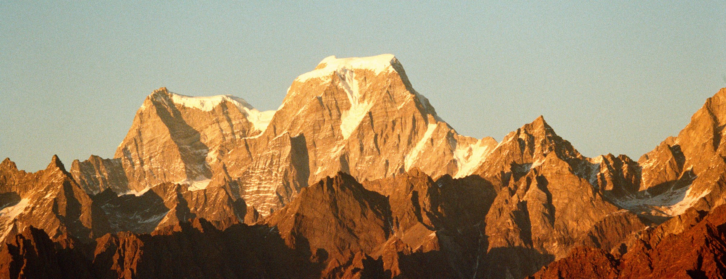 A high snow-capped massif standing above dark foreground ridges, lit amber by low sun under a pale, almost clear sky.